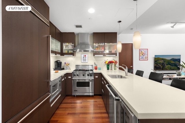 kitchen with built in appliances, hanging light fixtures, light countertops, wall chimney range hood, and a sink