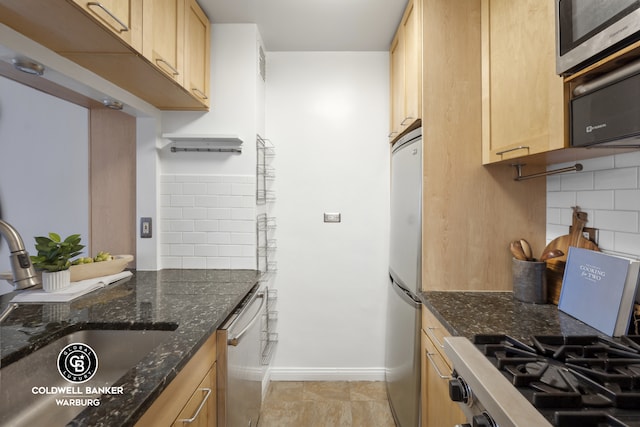 kitchen featuring a sink, appliances with stainless steel finishes, baseboards, light brown cabinets, and dark stone counters