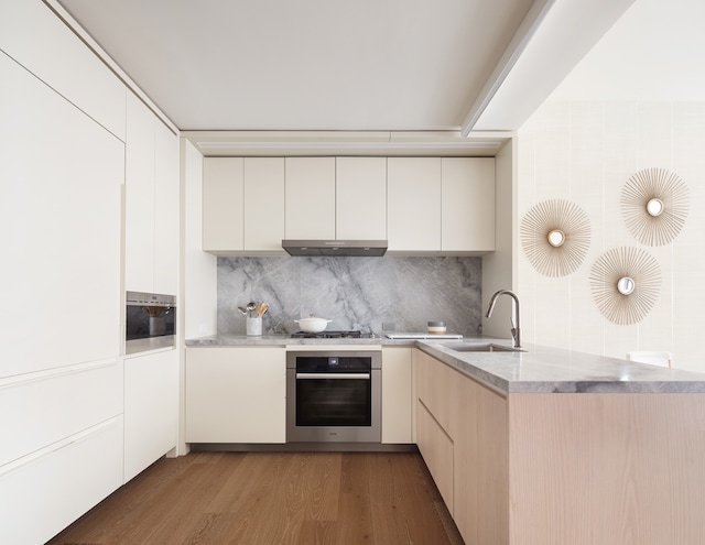 kitchen featuring stainless steel oven, a sink, modern cabinets, under cabinet range hood, and light wood-style floors