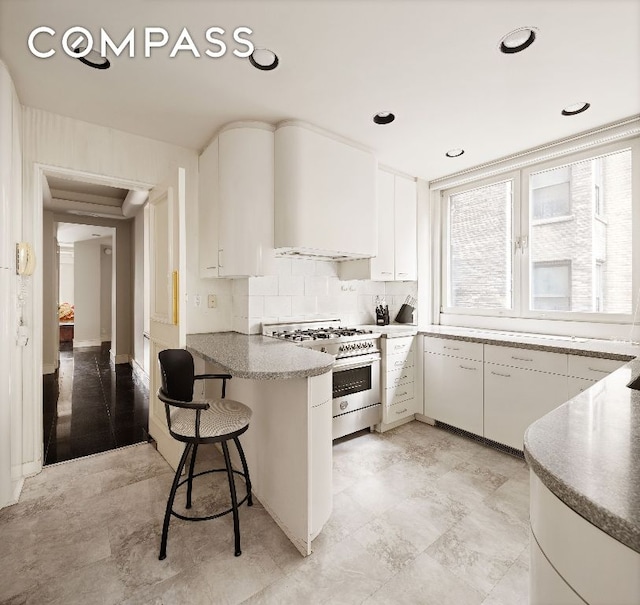 kitchen featuring stainless steel range, wall chimney exhaust hood, kitchen peninsula, white cabinets, and a breakfast bar area