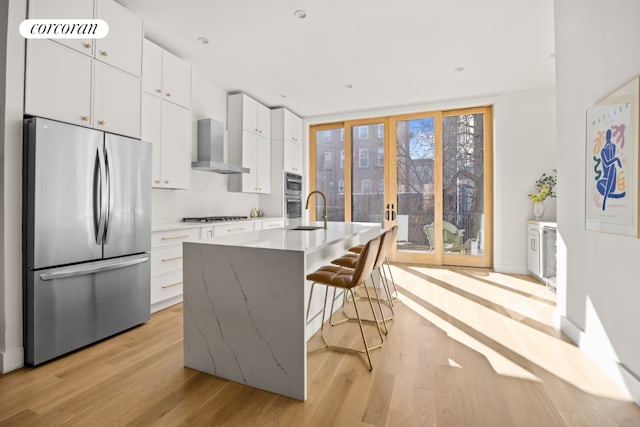 kitchen with a center island with sink, white cabinets, appliances with stainless steel finishes, wall chimney range hood, and a breakfast bar area