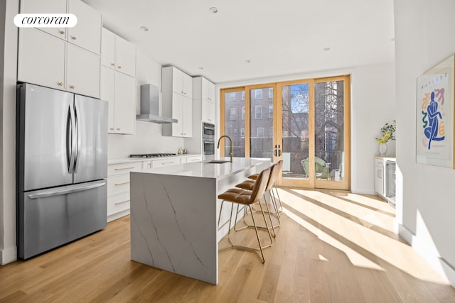 kitchen featuring a kitchen bar, light wood-style flooring, appliances with stainless steel finishes, white cabinets, and wall chimney range hood