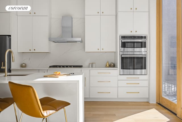 kitchen with a sink, appliances with stainless steel finishes, wall chimney exhaust hood, white cabinets, and light countertops