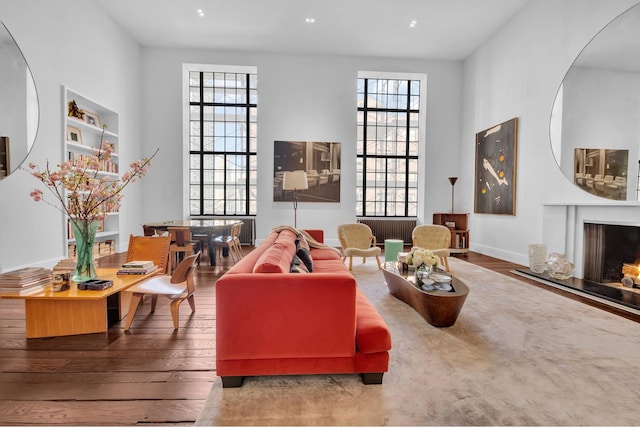 living area with built in shelves, a lit fireplace, a towering ceiling, and wood finished floors