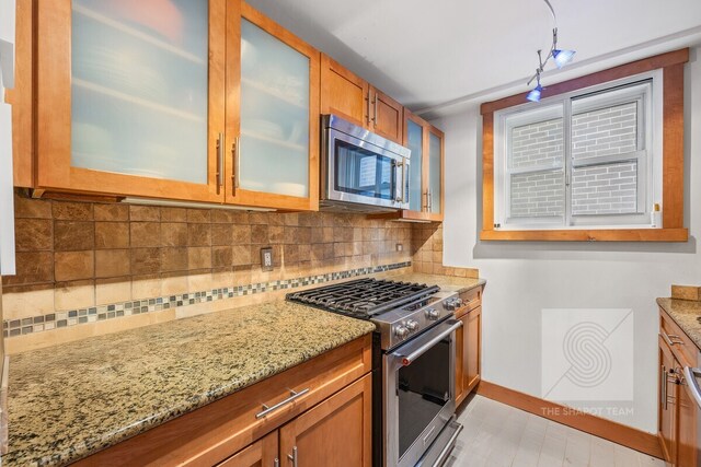 345 E 56th St Unit 21C, New York City, NY 10022 - kitchen featuring brown cabinets, stainless steel appliances, decorative backsplash, glass insert cabinets, and light stone countertops