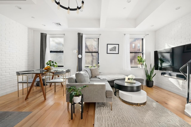 living room featuring brick wall, light wood-style flooring, a tray ceiling, and visible vents