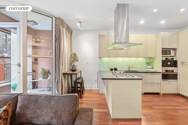 kitchen with light wood-style flooring, island exhaust hood, appliances with stainless steel finishes, dark countertops, and backsplash