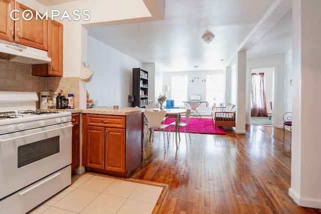 kitchen featuring under cabinet range hood, brown cabinetry, light countertops, and white range with gas cooktop