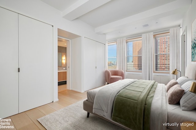 bedroom featuring two closets, beamed ceiling, light wood-style floors, and ensuite bathroom