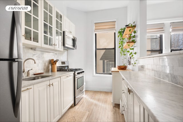 kitchen featuring appliances with stainless steel finishes, glass insert cabinets, white cabinetry, a sink, and light wood-type flooring