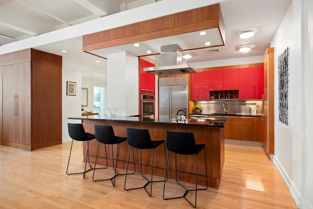 kitchen with light wood finished floors, a breakfast bar, island exhaust hood, stainless steel appliances, and dark countertops