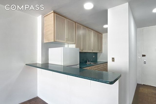 kitchen with dark countertops, freestanding refrigerator, light brown cabinets, a sink, and a peninsula