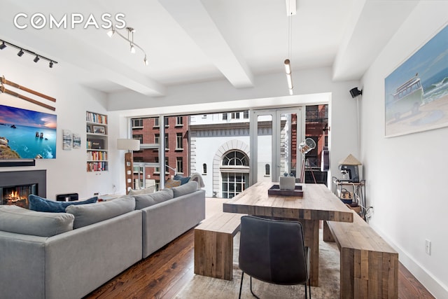 living area featuring dark wood-style floors, built in shelves, track lighting, a warm lit fireplace, and beamed ceiling