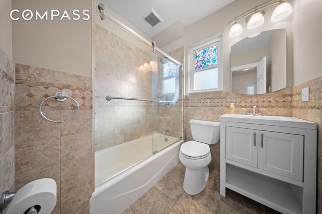 bathroom featuring toilet, shower / bath combination with glass door, visible vents, vanity, and tile walls