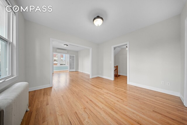 unfurnished room featuring radiator heating unit, baseboards, light wood-style flooring, and a wall mounted air conditioner