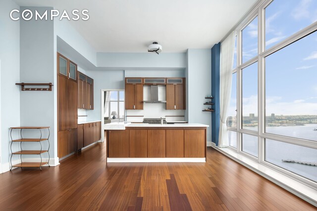 kitchen featuring dark wood-type flooring, baseboards, light countertops, wall chimney range hood, and glass insert cabinets