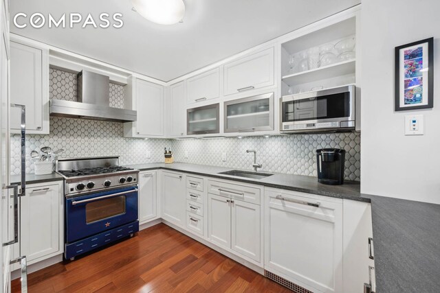 kitchen featuring dark countertops, appliances with stainless steel finishes, white cabinetry, a sink, and wall chimney range hood