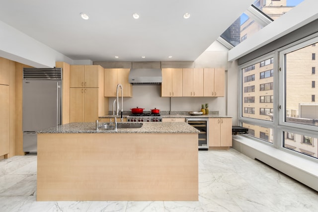 kitchen with marble finish floor, a kitchen island with sink, stainless steel built in refrigerator, wall chimney range hood, and light brown cabinets