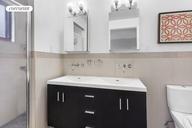 bathroom with toilet, tile walls, wainscoting, double vanity, and a stall shower