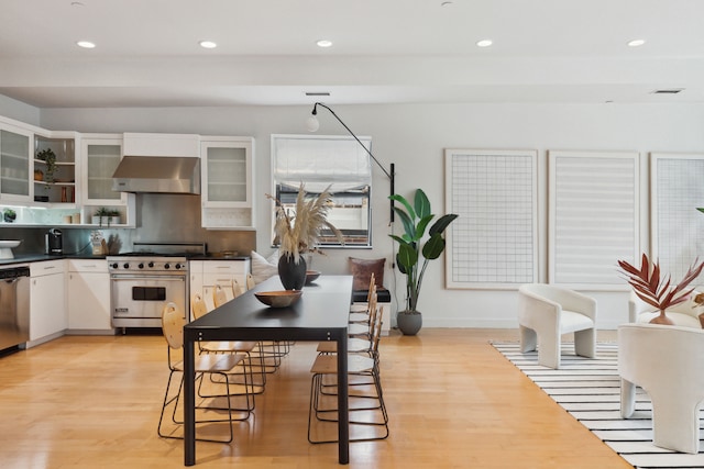 kitchen featuring white cabinets, dark countertops, glass insert cabinets, stainless steel appliances, and wall chimney range hood