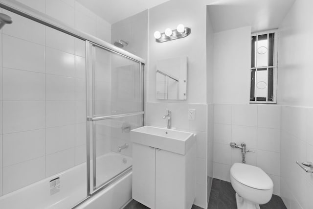 full bath featuring toilet, bath / shower combo with glass door, tile walls, and vanity