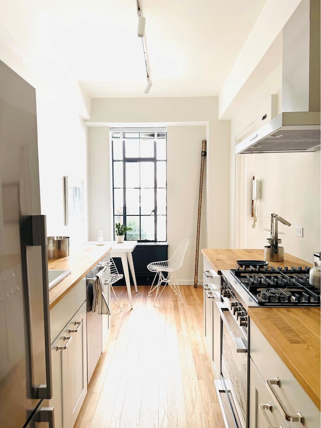 kitchen featuring rail lighting, appliances with stainless steel finishes, light wood-style floors, wood counters, and wall chimney exhaust hood