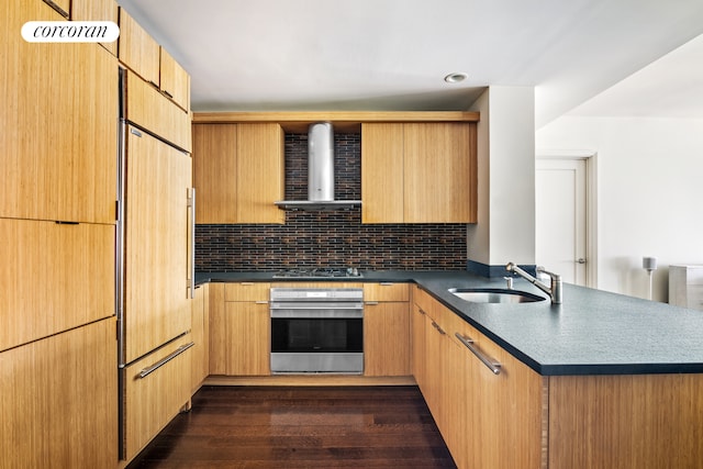 kitchen featuring stainless steel appliances, dark countertops, a sink, a peninsula, and wall chimney exhaust hood