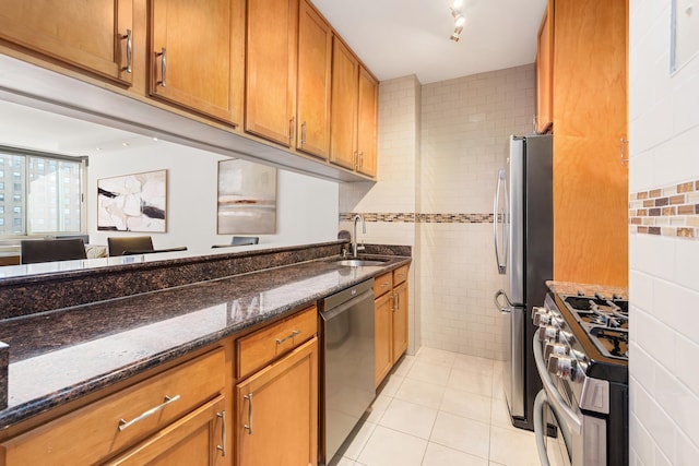 kitchen with a sink, tile walls, appliances with stainless steel finishes, dark stone counters, and brown cabinetry