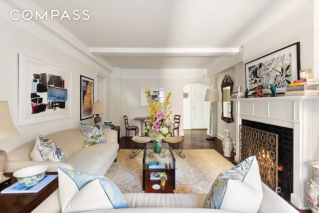 living room featuring arched walkways, beamed ceiling, a warm lit fireplace, and wood finished floors