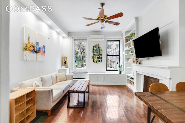 living area featuring built in features, a wall unit AC, a fireplace, a ceiling fan, and hardwood / wood-style flooring