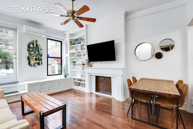 living area with built in features, a fireplace, a ceiling fan, wood finished floors, and a wall mounted air conditioner