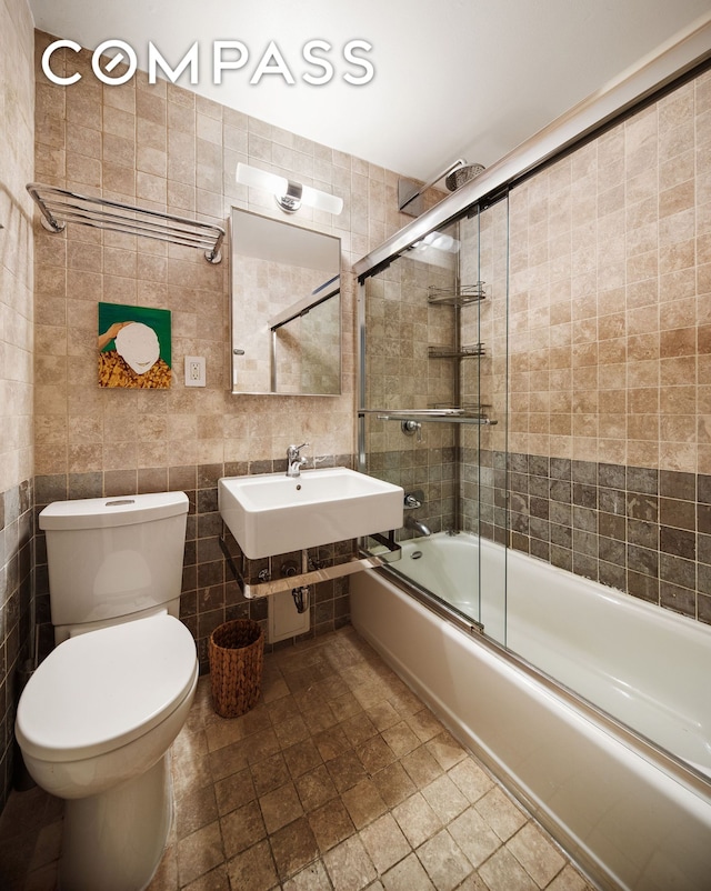 full bathroom featuring toilet, bath / shower combo with glass door, a sink, and tile walls