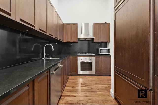 kitchen featuring brown cabinets, light wood finished floors, stainless steel appliances, a sink, and wall chimney range hood