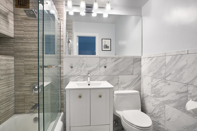 full bathroom featuring tile walls, toilet, shower / bath combination with glass door, vanity, and a wainscoted wall