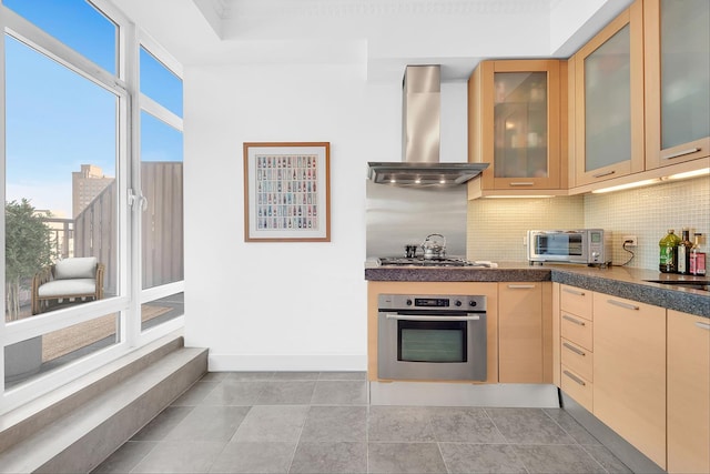 kitchen featuring a toaster, glass insert cabinets, appliances with stainless steel finishes, wall chimney range hood, and backsplash