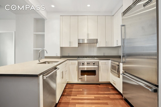 kitchen with built in appliances, under cabinet range hood, a peninsula, wood finished floors, and a sink