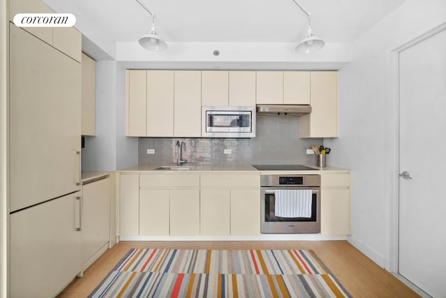 kitchen with stainless steel appliances, cream cabinetry, light countertops, under cabinet range hood, and a sink