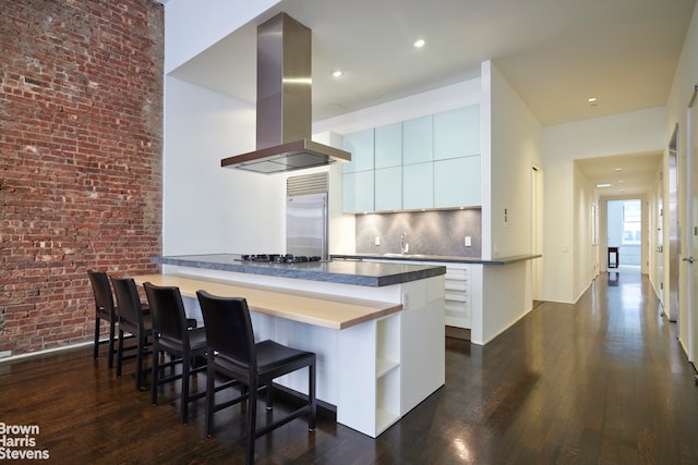 kitchen with island exhaust hood, dark wood-type flooring, appliances with stainless steel finishes, brick wall, and a kitchen bar