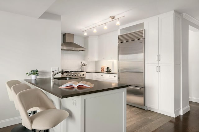 kitchen with stainless steel built in fridge, dark countertops, wood finished floors, white cabinets, and wall chimney range hood