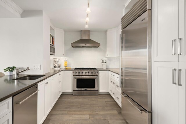 kitchen with built in appliances, wood finished floors, a sink, wall chimney range hood, and dark countertops