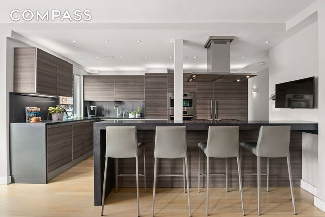 kitchen featuring double oven, dark countertops, island range hood, and modern cabinets