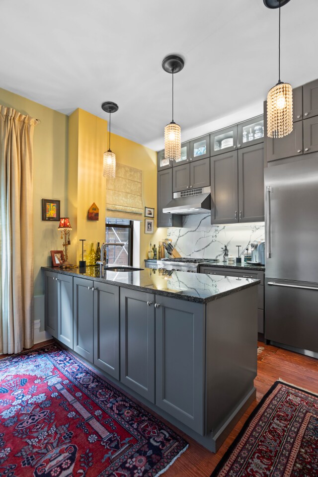 kitchen featuring dark wood-style flooring, dark stone countertops, a peninsula, under cabinet range hood, and stainless steel built in refrigerator