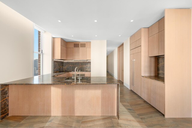 kitchen with dark stone counters, a peninsula, a sink, light brown cabinets, and backsplash