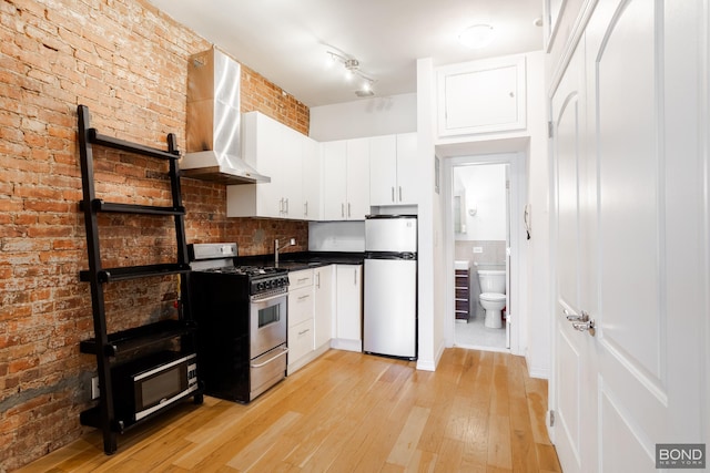 kitchen featuring gas range, wall chimney exhaust hood, freestanding refrigerator, light wood-style floors, and white cabinetry