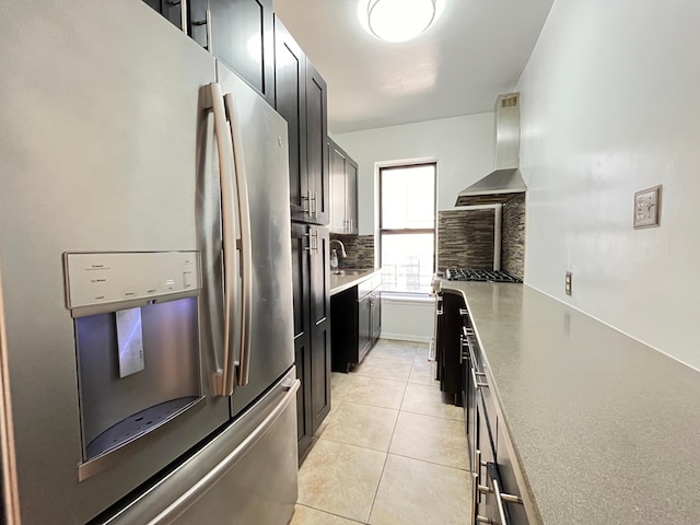 kitchen with light tile patterned floors, dark cabinets, a sink, appliances with stainless steel finishes, and wall chimney range hood