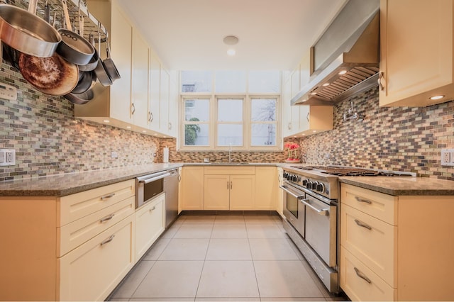 kitchen with a sink, stainless steel appliances, wall chimney exhaust hood, light tile patterned flooring, and decorative backsplash