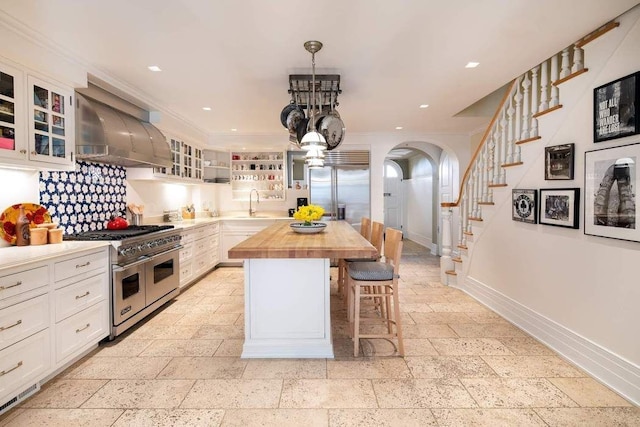 kitchen featuring wall chimney exhaust hood, high quality appliances, arched walkways, white cabinets, and butcher block counters