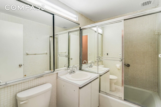 bathroom with visible vents, toilet, shower / bath combination with glass door, tile walls, and vanity