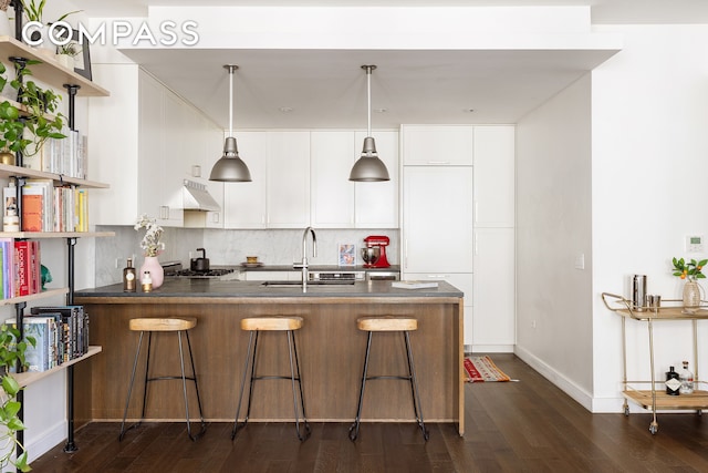 kitchen with open shelves, under cabinet range hood, a sink, dark countertops, and a peninsula