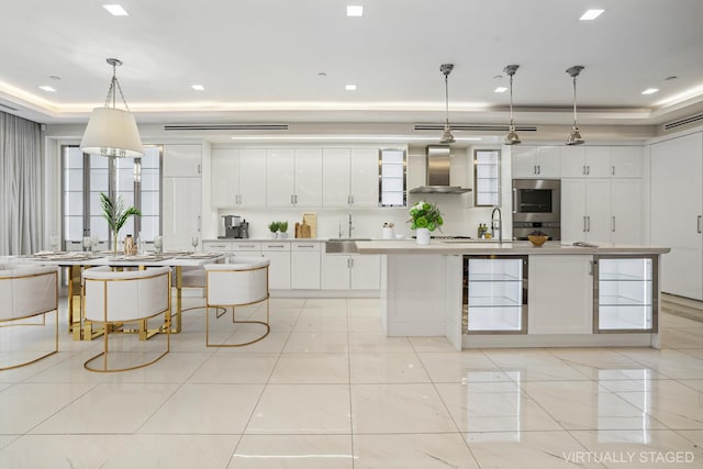 kitchen featuring stainless steel double oven, a sink, white cabinetry, wall chimney exhaust hood, and an island with sink